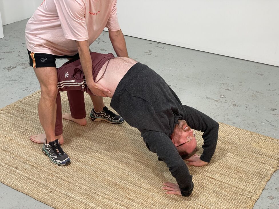 Nic Chilvers (left) helps Anthony (right) elevate his pelvis. Arching the body upward with assistance helps bring oxygen into Anthony’s lungs, while relieving fatigue and tension in the lower back, chest, and stomach. This gestural movement explores the notion of working with injury, identifying agency, and maintaining body mobility.