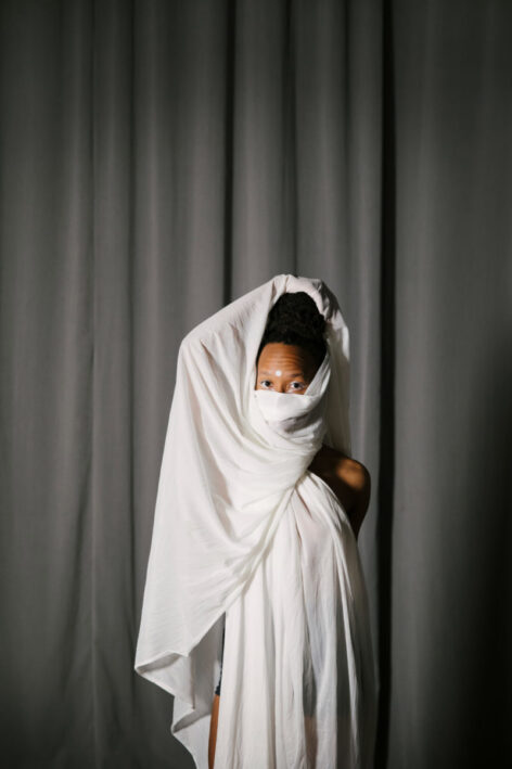  Portrait of Imani Rameses. She wears a long white dress and uses a white fabric to cover the bottom of her face. Her eyes look into the camera. Photo courtesy of the artist.