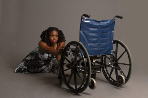 India Harville, African American cis woman with curly black locs in pigtails is crawling and reaching for her manual wheelchair with her right hand. She is wearing studded makeup jewelry on her face that mimics teardrops and is looking intensely into the camera. She is wearing a long sleeveless black and white tie-dye dress with a spider web-like pattern. Photo by Lexi.