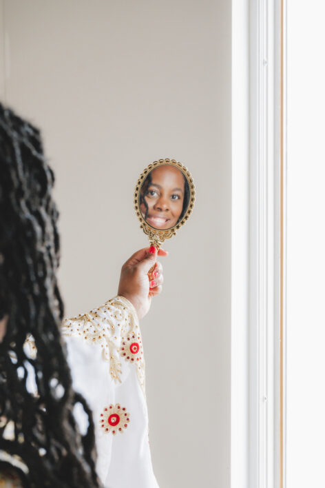 India Harville, Black, Disabled cis-gendered woman with long locs, holds an ornate, vintage-style hand mirror with a delicate gold frame. Reflected within the mirror is India’s smiling face. She wears a white garment adorned with intricate gold embroidery and red floral accents. Natural light streams in from a window.