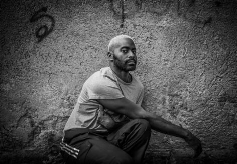 Black and white image of Jerron kneeling in front of a grey wall with loose graffiti staring into the camera. Image by Adrian McCourt.