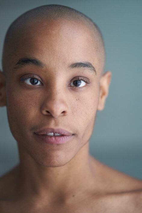 Headshot of Jasmine, a Black person with a shaved head, against a gray background. photo @Haym Heron