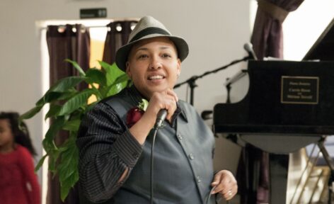 JJ Omelagah, a light-skinned black transgender person smiling with short faded black hair, wearing a gray and black fedora, gray and black long-sleeve collar shirt with a gray vest. They are singing to a crowd while holding a mic in their right hand and the cord in their left hand. Photo courtesy of Champion Fleming Design