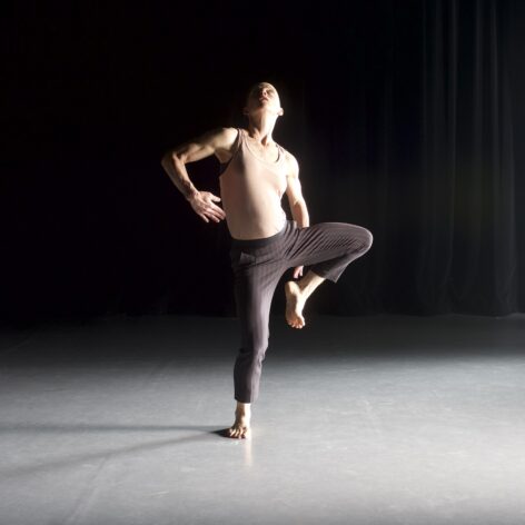 A dancer wearing a beige tank top and brown and purple pin stripped pants; on one leg while the other leg, foot, and knee are bent and curved; one elbow bent as the other hand reaches behind for the lifted foot. Chest lifted, back arched, gaze is at the ceiling. Photo by Jim Coleman.