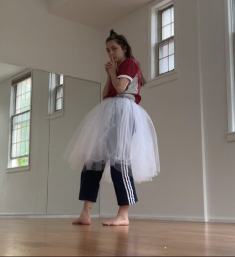 A photo of Julia, standing in a studio and looking into the camera. She is wearing a red top and track pants under a romantic tutu. She holds her hands by her mouth. Photo courtesy of the artist.