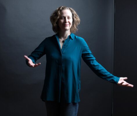 Julie Mayo photo by Ebru Yildez. Photo of Julie smiling, stands with soft arms wide open in a blue long sleeve top.