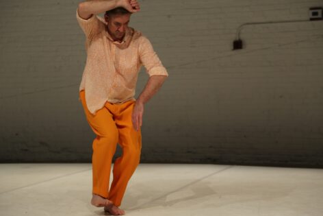 A photo of Jon Kinzel. He wears a light beige shirt with orange pants. One arm is up and he is looking down, while one foot is on the white ground. The walls are white brick. photo by Brian Rogers