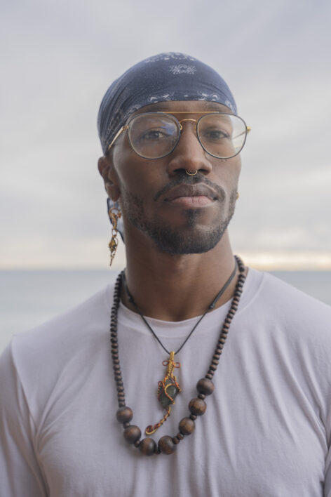 A portrait of j. bouey in a white tshirt, glasses, and bandana. they are adorned in various pieces of jewelry, a septum piercing, an earring in one ear, and two necklaces.