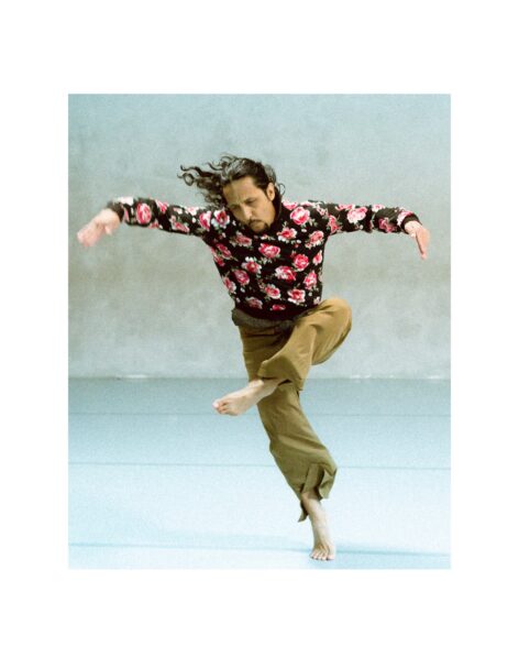 A person with long dark hair wears a black floral shirt and tan pants while dancing with one leg bent and the other extended. Luis A. Lara Malvacías photo by Arnaud Beelen.
