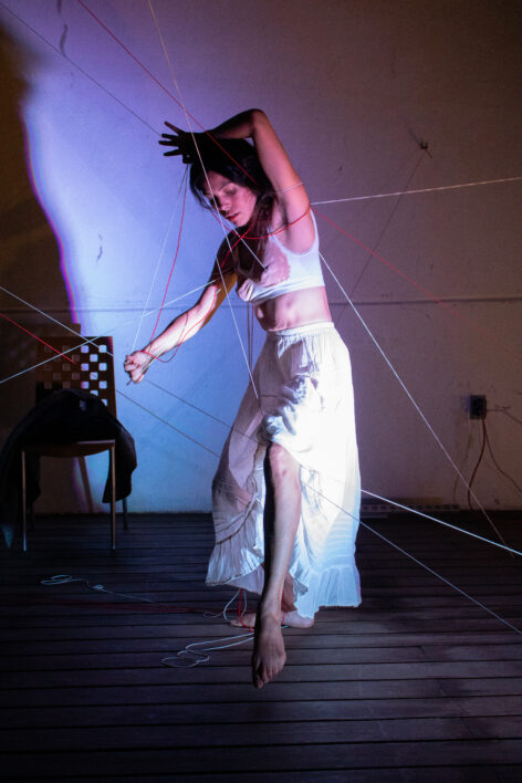 Maira Duarte is performing in front of a white wall, at night. She's wearing a white tank top and a white long skirt. With one arm above her head and one at her side she moves through strings that cross the space at different angles. Photo by Micelio Media.