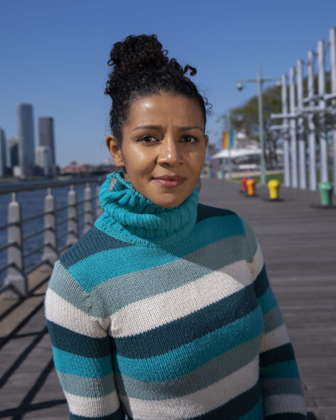 A photo of Maria Bauman posing for a headshot in a pier. She wears a blue and white stripped turtle neck sweater and wears her dark curly hair in a high bun. She looks into the camera with a warm expression. Photo by Lola Flash