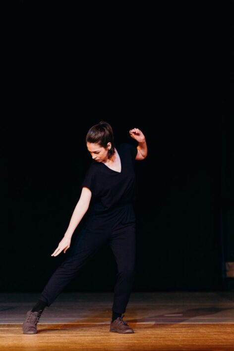 Mariam wears a blue jumpsuit and charcoal sneakers in a darkened theater. While dancing, she bends and looks downward to the side, her leg and arm are both extended. Photo by Sarah Berman.