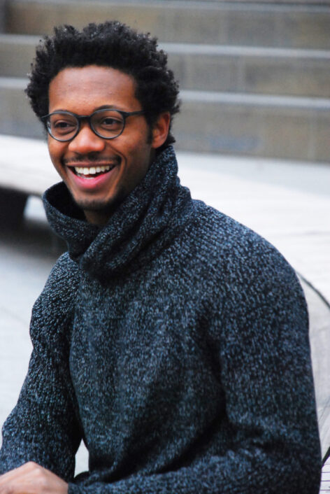 Kyle Marshall poses for a portrait. He is smiling and looking away from the camera. He wears a dark gray knitted turtle neck sweater and dark rimmed glasses. Photo by Norbert De La Cruz III.