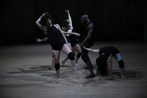 A grouping of 5 people caught mid action: Talya leans right arm over head leg slightly raised, Paul in profile legs apart sunken head slightly down, sam touching his hand with hers the other hand on the floor legs apart head over, laurel mid-stride arches sideways towards the us, Rainey is hidden by the rest of the group. Their shadows a jumble on the wooden floor. All wear minimal black outfits and knee pads, sam has on light blue stockings. Photo by Paula Court.