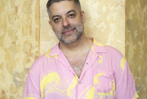 A cis, Latinx man with short dark hair and a greying beard looks directly at the camera with a slight smile. He is wearing a pink shirt with yellow jellyfish print and is photographed against a yellow marbled background. Photo by Chloe Cusimano.