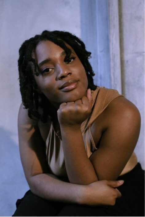 Portrait of Monica Nyenkan. She is seated and leaning forward with one arm resting on her land the other supporting her head. She is looking into the camera. She wears a light brown top and her hair is in locs. Photo by Jakob Tillmann.
