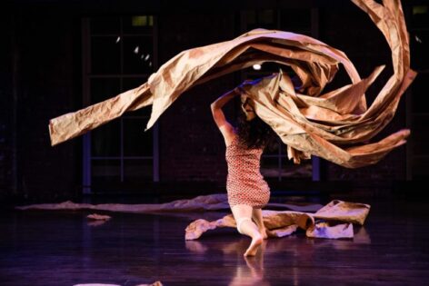 Photo of Nami Yamamoto performing. She is on stage lunging away from the camera. She waves a large bundle mede up of long strips of brown paper over her head. She wears a short patterned dress. Photo by Scott Shaw