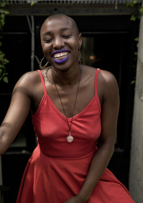 Mid-body portrait of Nana Chinara with a vibrant and full smile and eyes closed. Nana wears big rounded earrings, a pendant, a colorful tank top, and bright purple lipstick. Photo by Kalyn Jacobs.