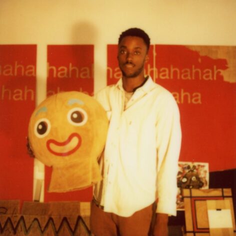 Nile stands in front of a red painting that reads 'ha ah ha'. He is wearing a white button up shirt and holding the headpiece of gingerbread man costume.