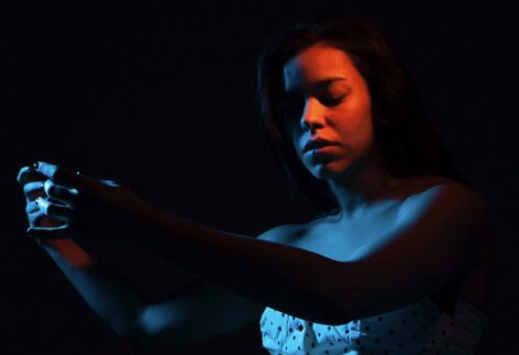 Portrait of Nora Alami looking downward with arms extended forward. Her body is lit in a blue light and her face is lit in amber. She is wearing a black and white polka dot top. Photo by Haim Machi Versano 