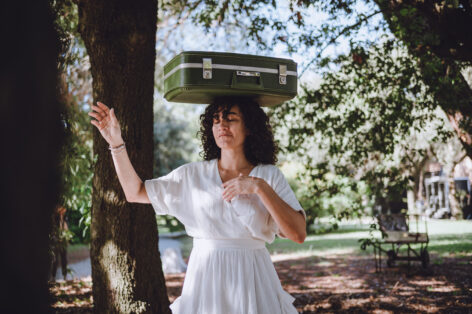 An image of Paloma McGregor, a sand colored Black woman standing in a white dress with one hand near her heart and her other hand in the air. She balances a suitcase on her head. There are trees and a light blue sky surrounding her. Photo courtesy of the artist.