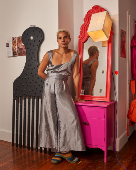 Paris Alexander, light-skinned mixed Black queer person, stands contrapposto in their bedroom looking directly toward the camera, one arm behind their back, one leaning on a bright pink dresser. They are wearing silver high-waisted pants with flaring wide-legs and a matching tank top cinched at the waist. A black pick comb as large as Paris and adorned in large sparkles rests against the wall next to them. Photo courtesy of the artist.