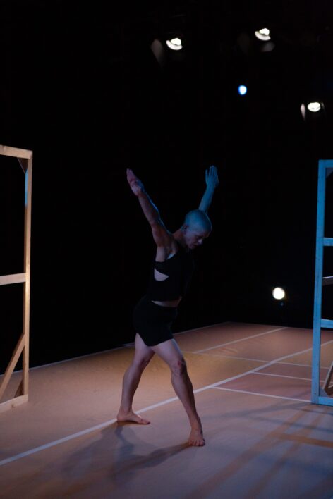 a white woman with a shaved head wearing black shorts and a black tank top in dim lighting, arms lifted overhead, head looking down, leans forward onto one leg. Photo by Julia Discenza.