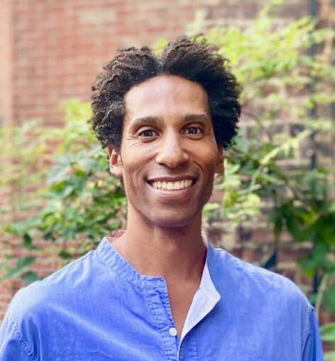 A photot of Stacy Matthew Spence smiling at the camera. He has short afro hair and wears a blue top. Photo by Michael Argilla