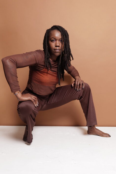 Image is of dance CocoMotion a black woman with long dark brown locs draping her face. Wearing a brown sheer shirt, orange bra and brown pants. Brown backdrop. Photo by Qlick Photography