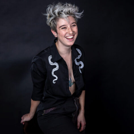 RK, a white femme presenting person with short bleached white hair, sits in front of a black background. They are leaning forward laughing. Their black shirt is open showing a glimpse of a green hummingbird tattoo on their torso. Photo taken by Lourdes Severny