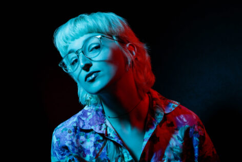 A headshot of Raychel Ceciro, a white person with white hair and glasses in pink and blue lighting. They are wearing a pink floral collared shirt. Photo By Sorcha Augustine.