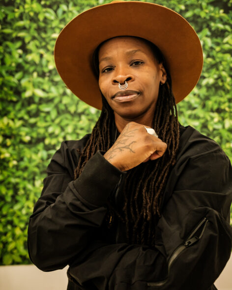 Sage, a dapper dark skinned Black transgender artist faces the camera with a sly smirk. They wear a wide brimmed brown hat on their head. They hair is in locs and they hold their arm over their torso, hand near their chin in a relaxed fist. Photo by Ryan Sandell.
