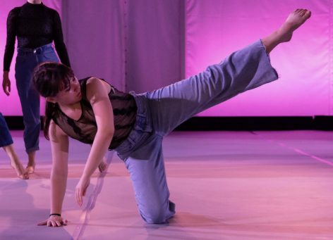 Hannah is wearing jeans and a black lace tank top. She is in motion, with her right hand and right knee planted on the ground, and her left leg raised above her to the side and her left arm draped in front of her. The stage lighting is hot pink. Photo by Jamie Kraus.