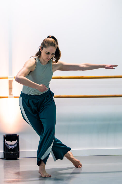 White woman with brown hair in green pants and shirt performing in a dance studio. Her right arm is bent over and left arm is lengthened out to the side. She is looking down and her left leg is bent in front of her right leg, which is lengthened behind her. Photo by John Eng.