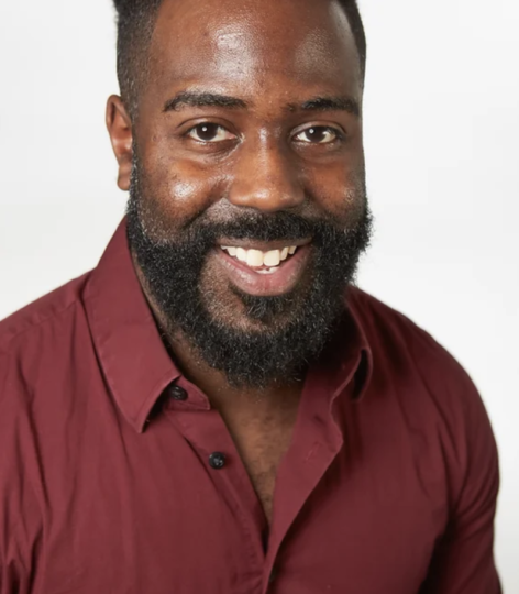 Headshot of Shawn Rawls smiling into the camera. He has a beard and wears a wine colored button up top. Photo courtesy of the artist. 