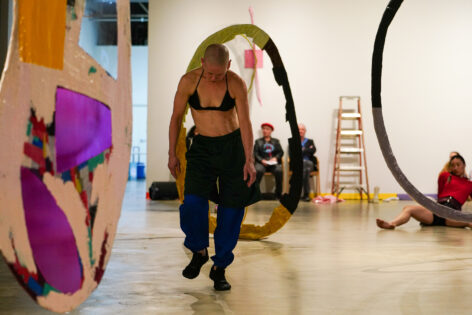 A white woman with a shaved head, wearing a bra, a cape, pants, and black running shoes, walking and looking downwards in a dimly lit museum gallery. Jennifer performs in Listeners by Beth Gill. Photo by Sidney Chansamone
