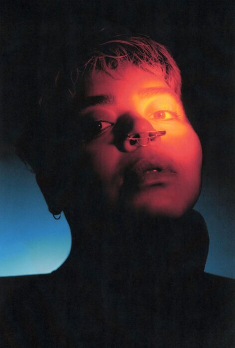 stefa marin alarcon gazes toward the camera with intent softness as a pool of warm orange and red light illuminates their left eye and dissipates across their face. Their nose pierced with a safety pin and a septum ring while the shadow of their ear discloses a small round earring. They are lit from behind with a small blue light deep in the image’s background, most of their head and collar left in dark silhouette. Photo courtesy of the artist.
