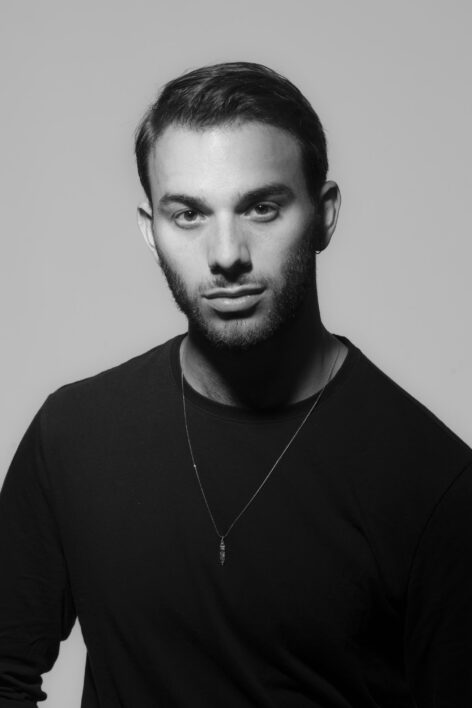 A black and white portrait of a white person wearing a black long-sleeved shirt, a thin gold necklace with a vertical pendant. They have short brown hair, a brown beard, and are staring into the camera's lens with a serious, but cunning look. Photo by Tsi Lang.