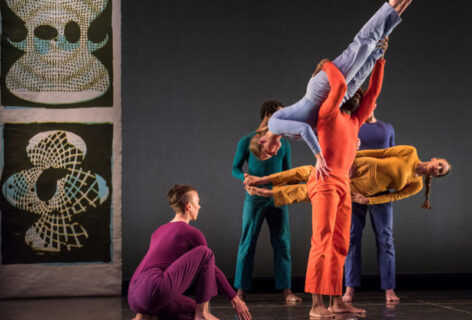 Photo of several dancers from the Trisha Brown Dance Company Performing. They wear bright monochrome costumes each in a different color. One dancer crouches down. Two dancers are lifted. Photo courtesy of the Trisha Brown Dance Company.