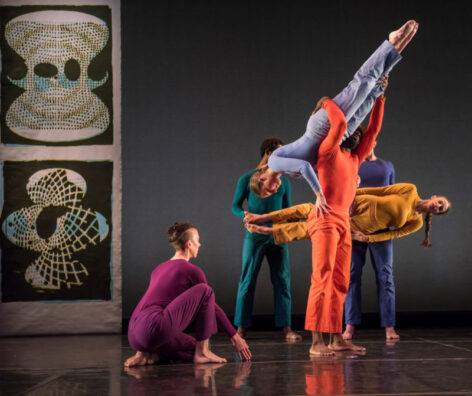 Photo of several dancers from the Trisha Brown Dance Company Performing. They wear bright monochrome costumes each in a different color. One dancer crouches down. Two dancers are lifted. Photo courtesy of the Trisha Brown Dance Company.