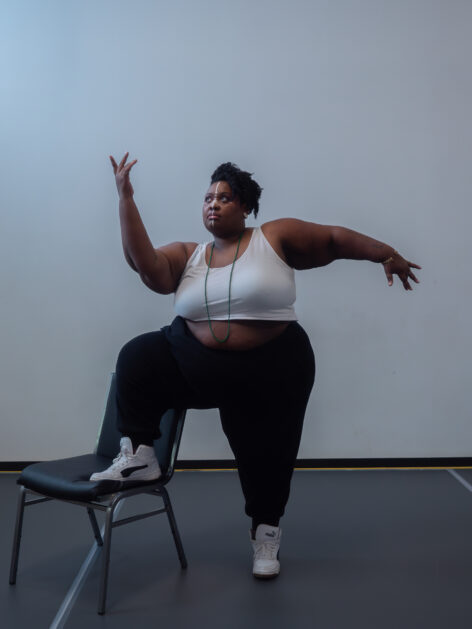 A fat Black genderqueer person standing with one of their legs on a chair. One arm is bent at the elbow with the fingers gesturing in the air, and the other arm is extended away from his body. They are wearing a white top, black pants, white and black sneakers.