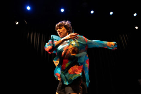 Performance photo of Tatyana Tenenbaum. She is dancing swaying her arms. She wears a blue and orange colorful top and has dark brown hair. Photo by Maria Baranova.