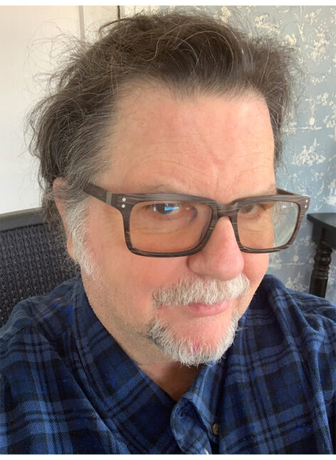 A photo of Tere O'Connor. He has a short white mustache and beard and wears a plaid blue top. He wears dark brown glasses and looks into the camera with a neutral expression. Photo courtesy of the artist.