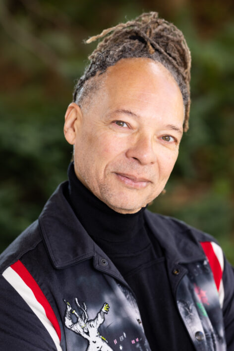 A beige man wearing a black turtleneck shirt sits for a portrait, and tilts his head with green eyes towards the viewer, his head crowned by reddish dreadlocks and a 'mona lisa' smile. Photo courtesy the artist.