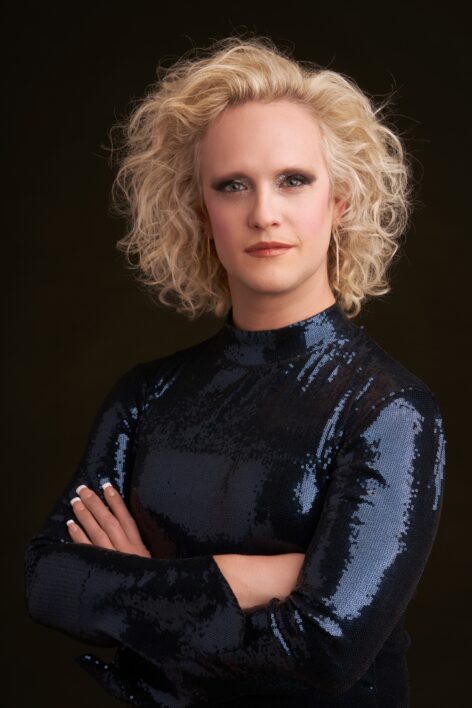 Tyler Ashley poses with arms crosses wearing a dark blue sequinned long sleeve top. Short Blonde wavy hair frames Tyler's face. Tyler looks into the camera with a neutral expression. Photo by Timothy Greenfield Sanders.