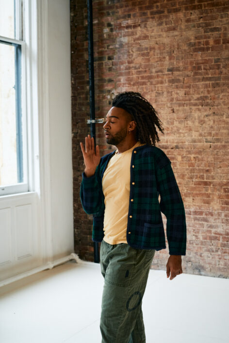 Wendell faces the right side of his room with his eyes closed and right hand paused in front of him. He is wearing a pale yellow shirt, and blue and green cardigan, and green pants. Photo by Tre Crews.