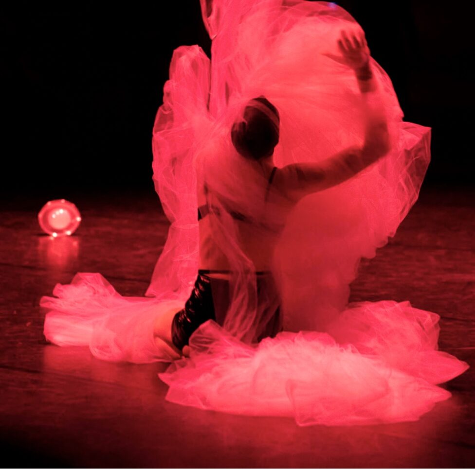 A kneeling person is shrouded in fabric lit pink by a light
