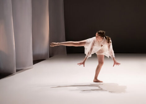 Photo of a dancer in performance. The dancer is balancing on one leg while the other reaches to the side. Their torso is bent over the standing leg while their arms reach straight down. The dancer looks towards the gesture leg and wears an all white costume. Photo by Derek Fowles.