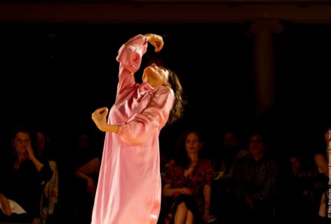 Darian Faïn performs wearing a shiny rose pink dress. Her arms are in motion scooping upwards before her while her torso and head reach diagonally back into the space. In the background the audience watches attentively. Photo by Alex Escalante.