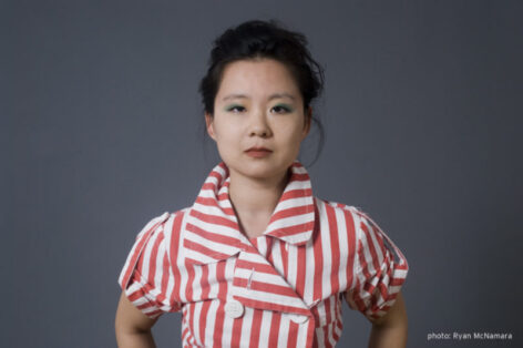 Standing before a grey background with shoulders slightly extended, Sam wears a red and white striped buttoned jacket with short, puffed sleeves and a statement collar. Photo courtesy of the artist. 